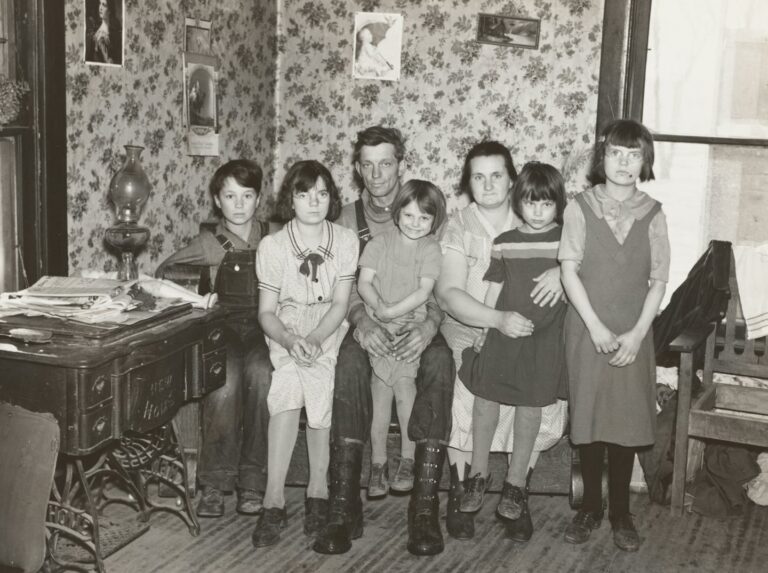 A family of eight posed together in a wallpapered room, the children in front of their parents.
